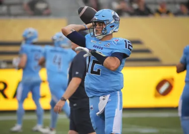 A football player in a light blue uniform with the number 12 prepares to throw the ball during a game.